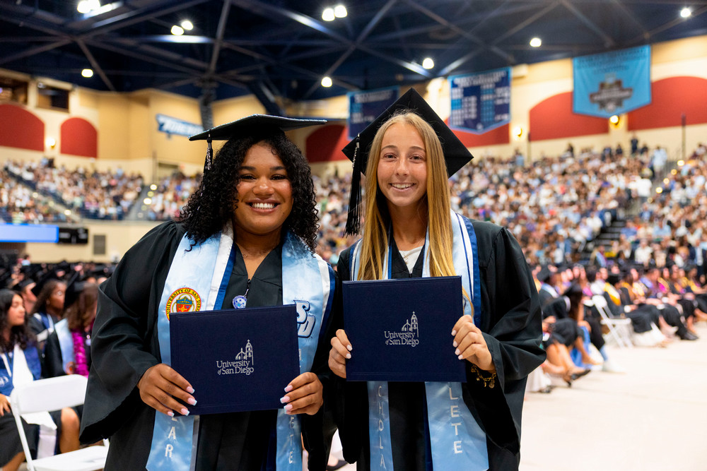 2 female students wearing graduation caps and robes holding up their diplomas
