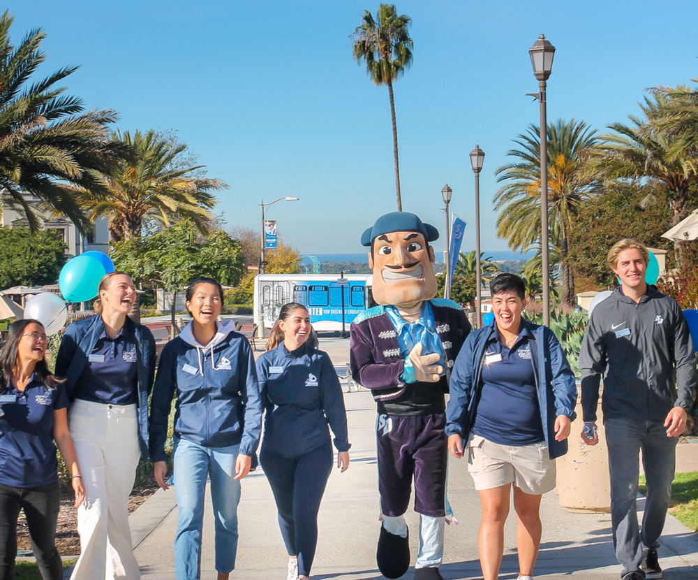 Photo of students wearing blue polos walking besides mascot on campus