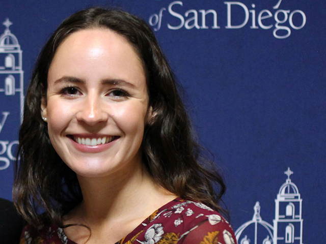 Student wearing red patterned dress smiling in front of blue USD backdrop