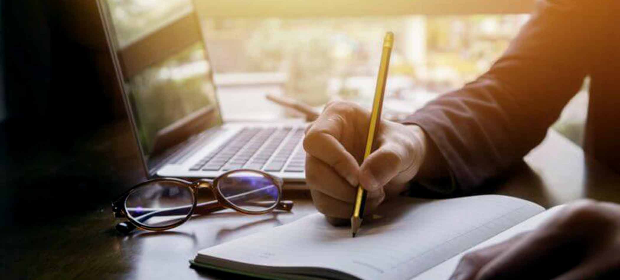 A person writing in a notepad with a laptop and glasses nearby