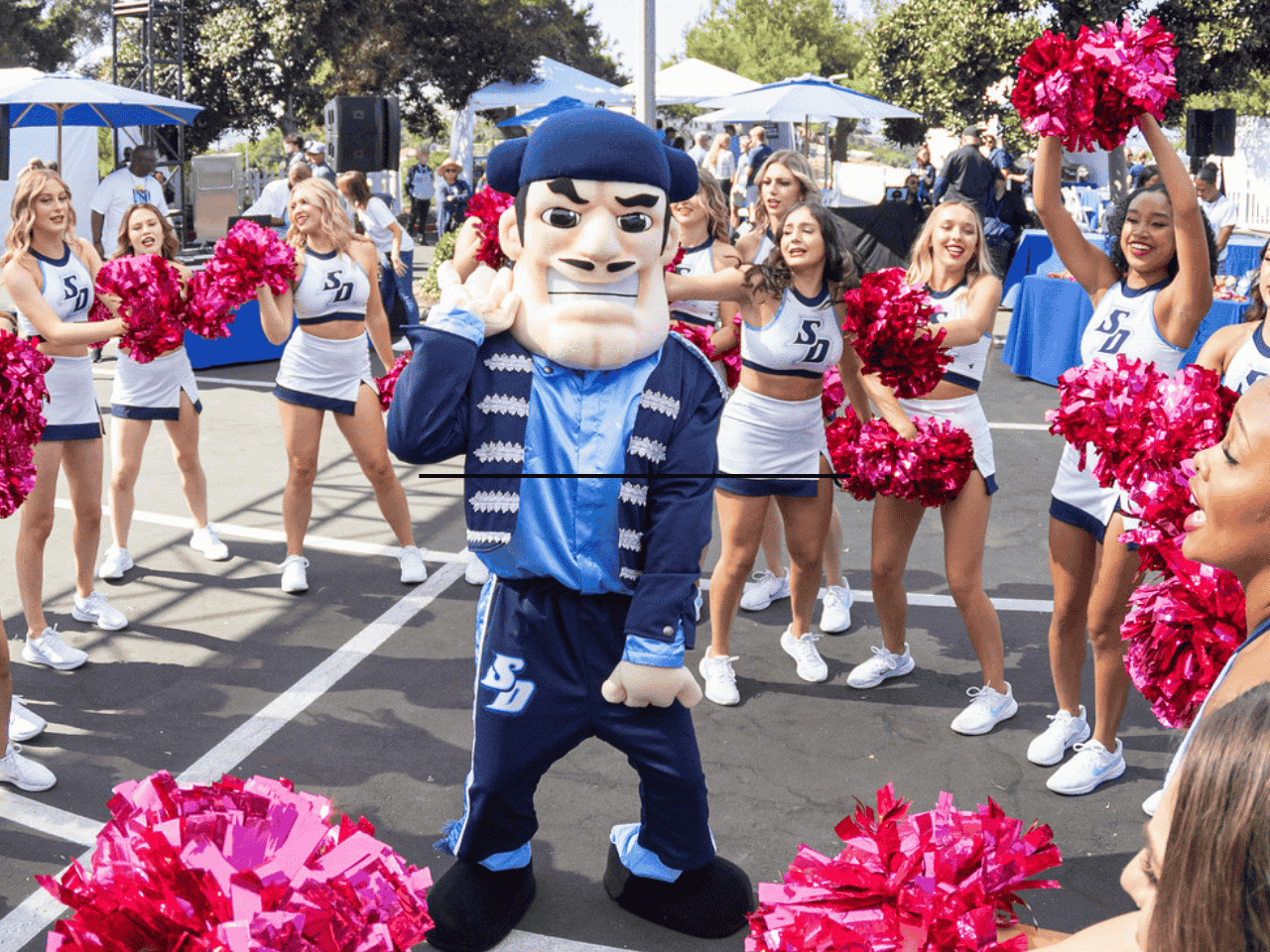 USD Mascot Diego with his hand up to his ear surrounded by cheerleaders.