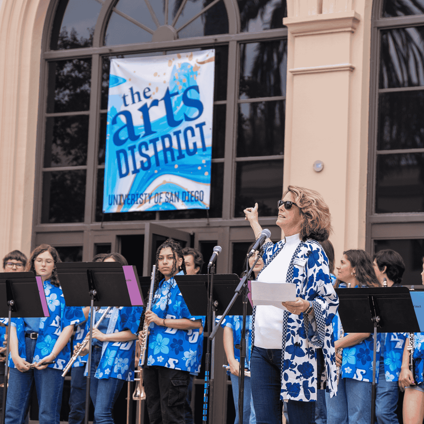 Dean Noelle Norton in front of an Arts District signwith a student band in the background.