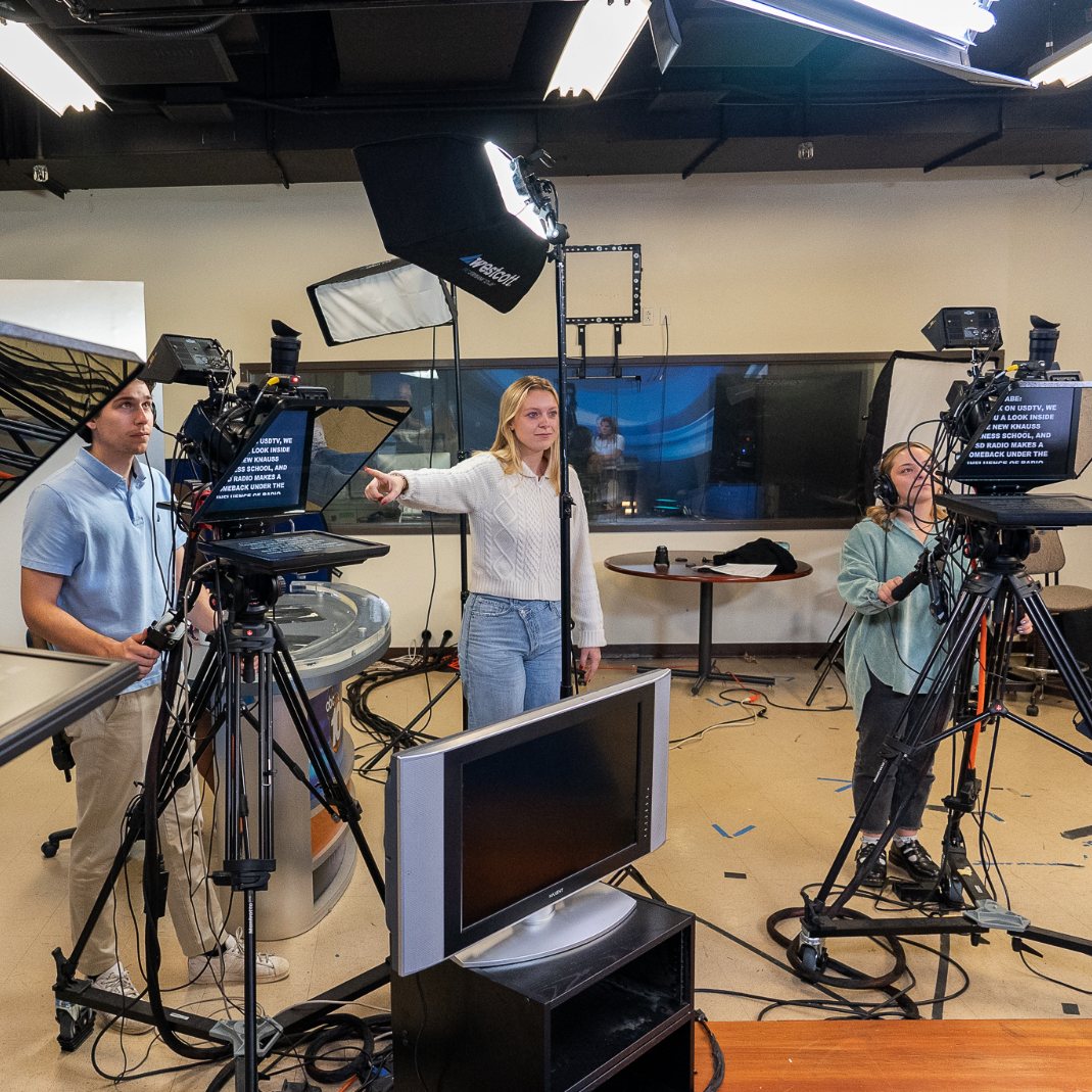 Students in a news broadcast studio working equipment during a television broadcast.