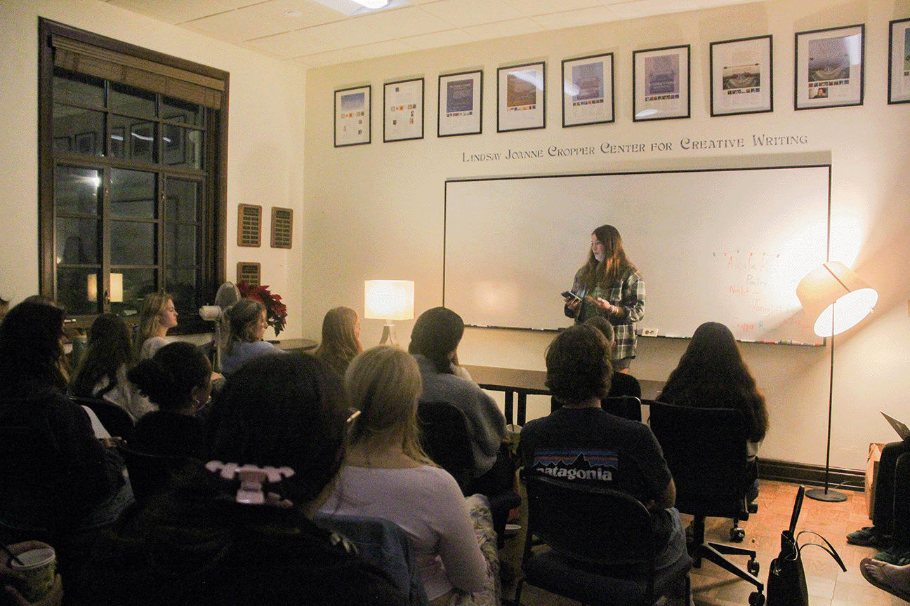 Students watch a person reading at the front of a dimly lit classroom.