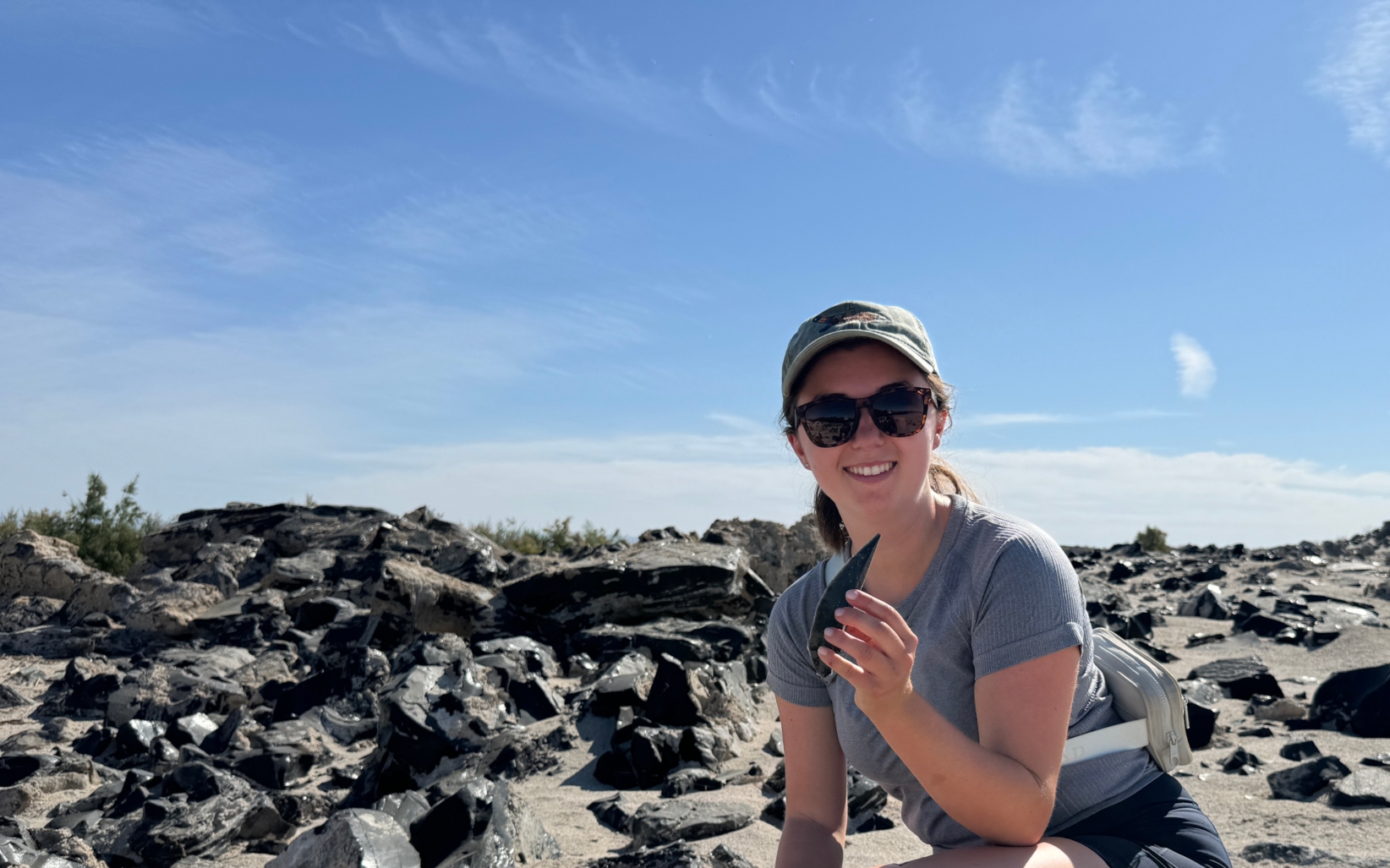 A student kneels on a rocky desert landscape, smiling while holding a piece of stone during fieldwork.