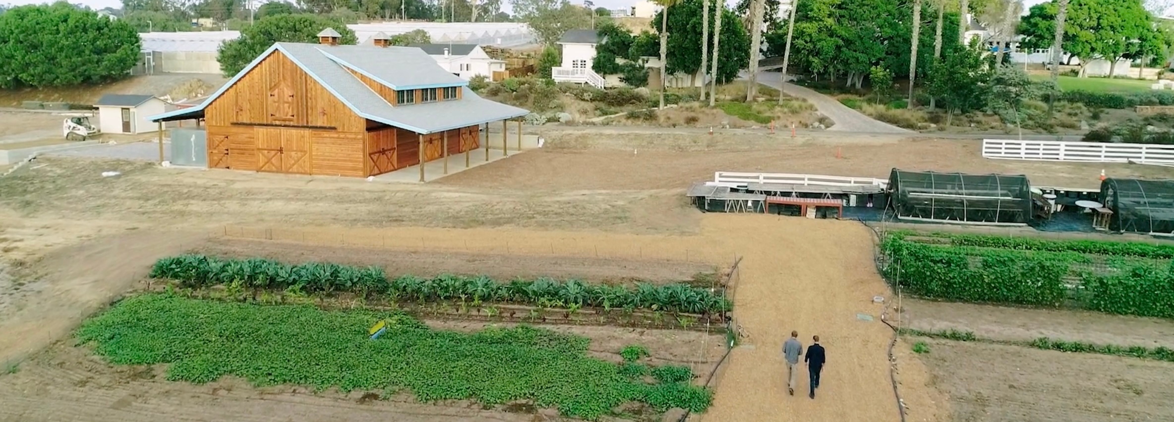 A drone shot of the a farm with a brown barn, a greenhouse, a green field and two people walking on on a path.