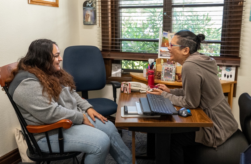 student sitting in office chair while talking to professor behind desk