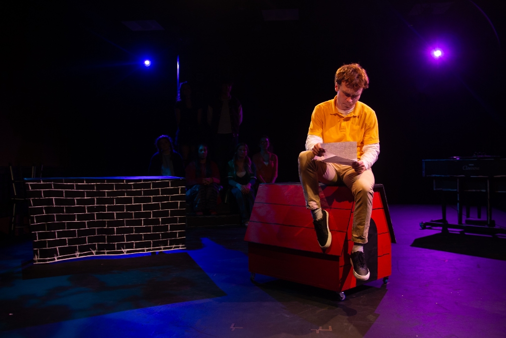 Male actor in a yellow shirt sits on a red dog house, reading a letter. The cast of characters sit grouped behind him, silhouetted by the lights.