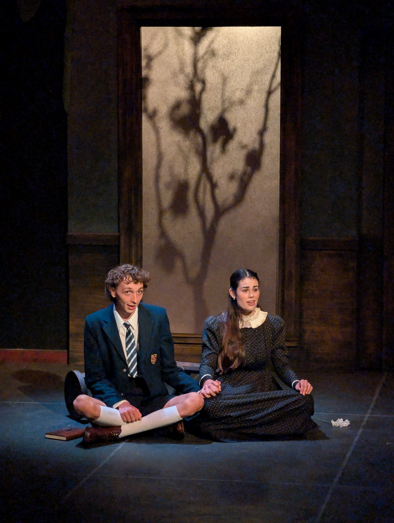A male actor and female actor sit cross-legged on the floor side by side holding hands, dressed in 19th century German school uniforms. Behind them is the silhouette of a mostly-barren tree. The actors look hopefully off into the distance.