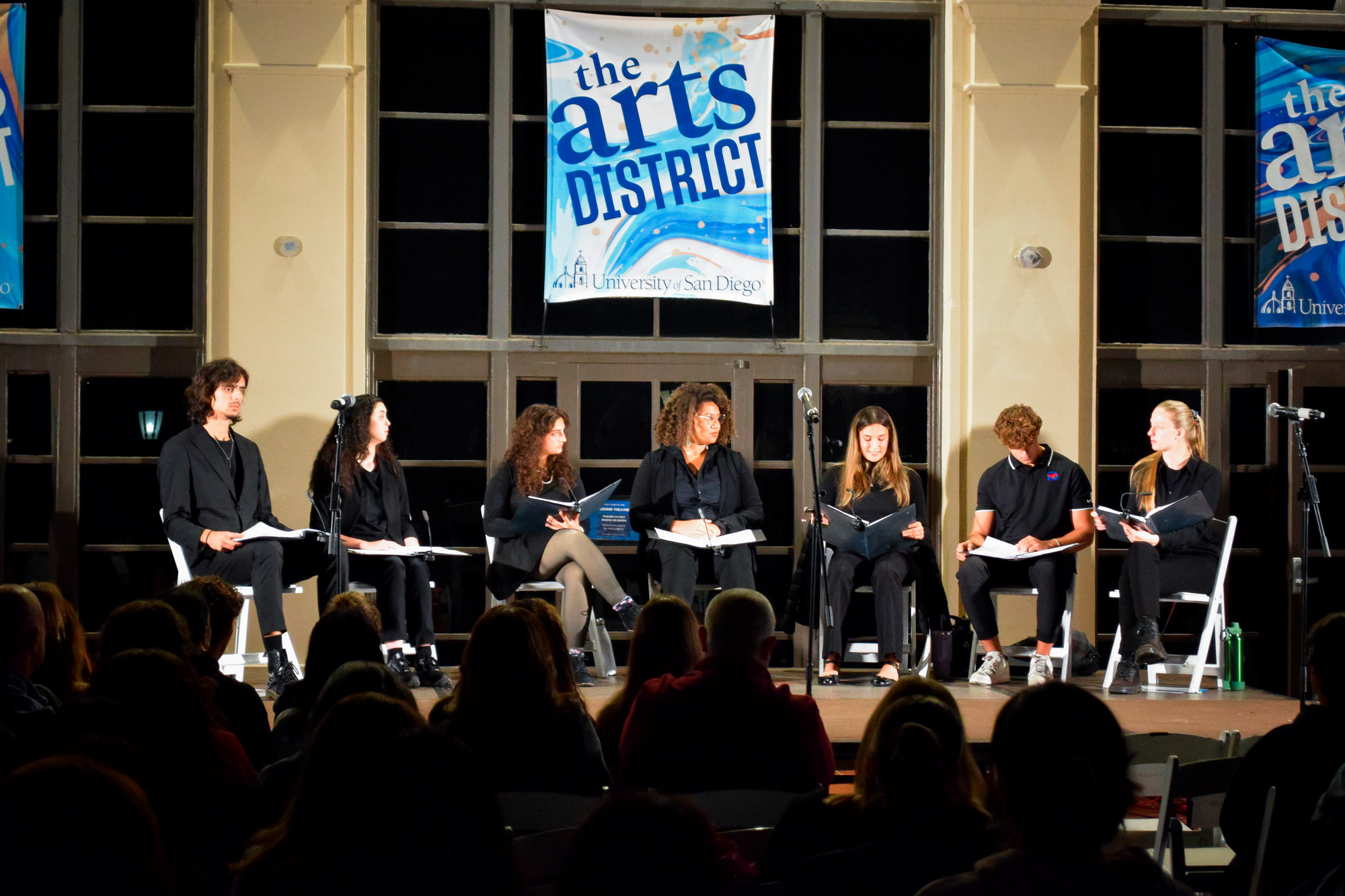Seven actors seated next to each other on a stage facing an audience during a staged reading of Twilight: Los Angeles 1992.