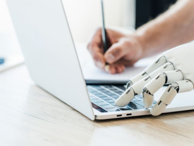 A human hand and robot hand typing on a laptop.