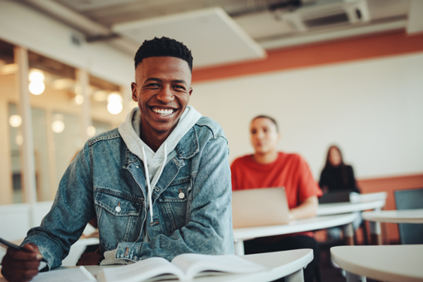 A student, sitting at a desk with an open book, looks into the camera smiling.