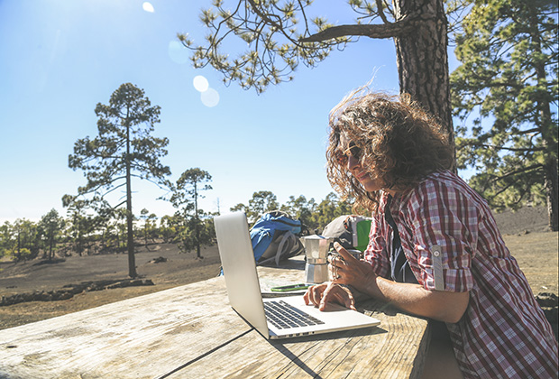 light-skinned female presenting person sitting at a picnic table with a laptop. The sun is shining and there are trees surrounding.