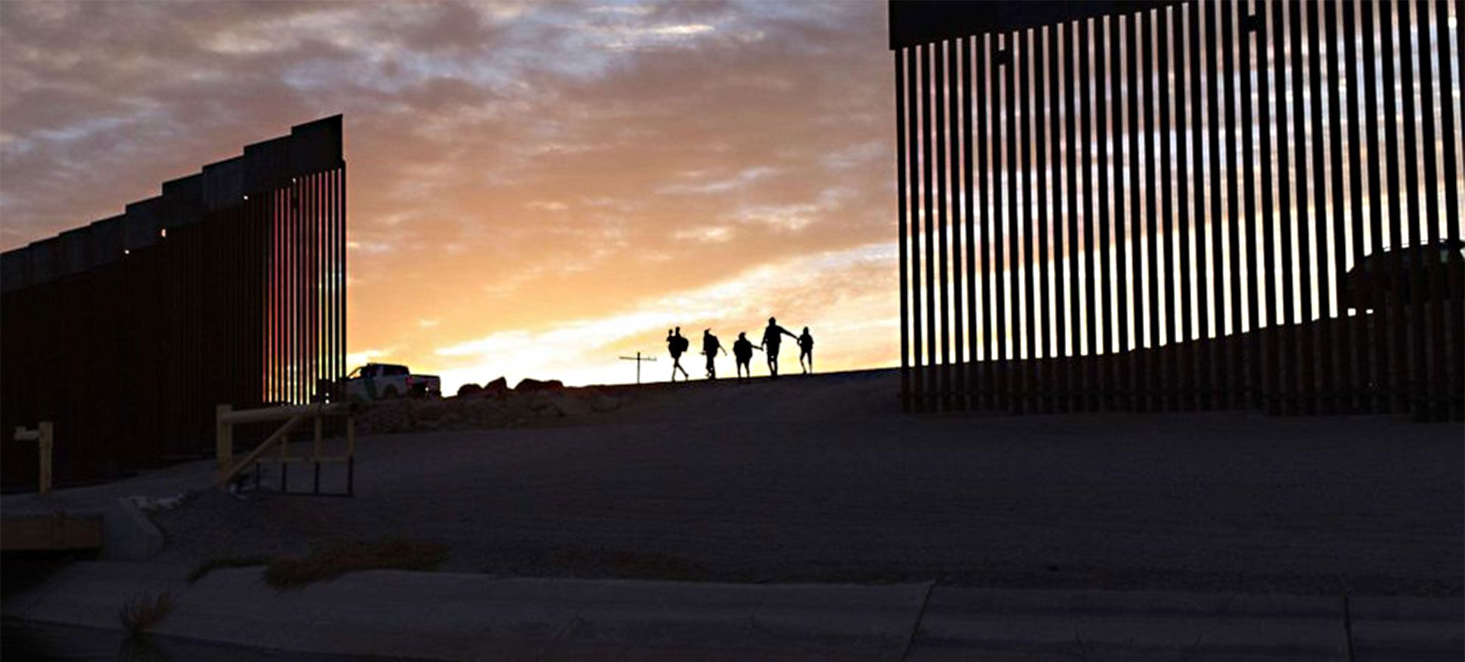 U.S. Mexico border wall at sunset with silhouettes of people walking along it