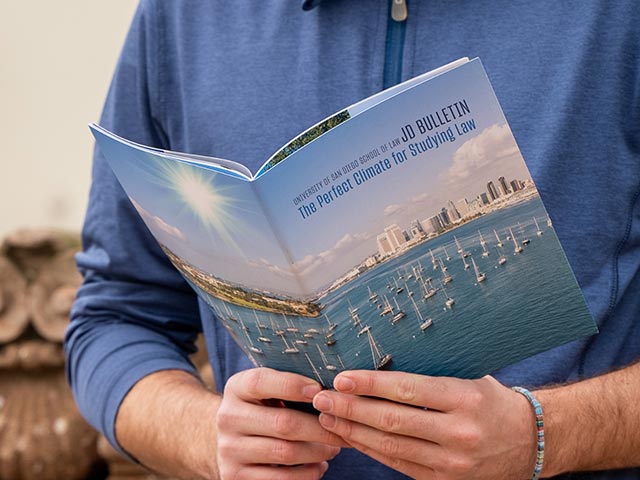 Someone reading a pamphlet. The cover reads "University of San Diego School of Law JD Bulletin. The Perfect Climate for Studying Law."