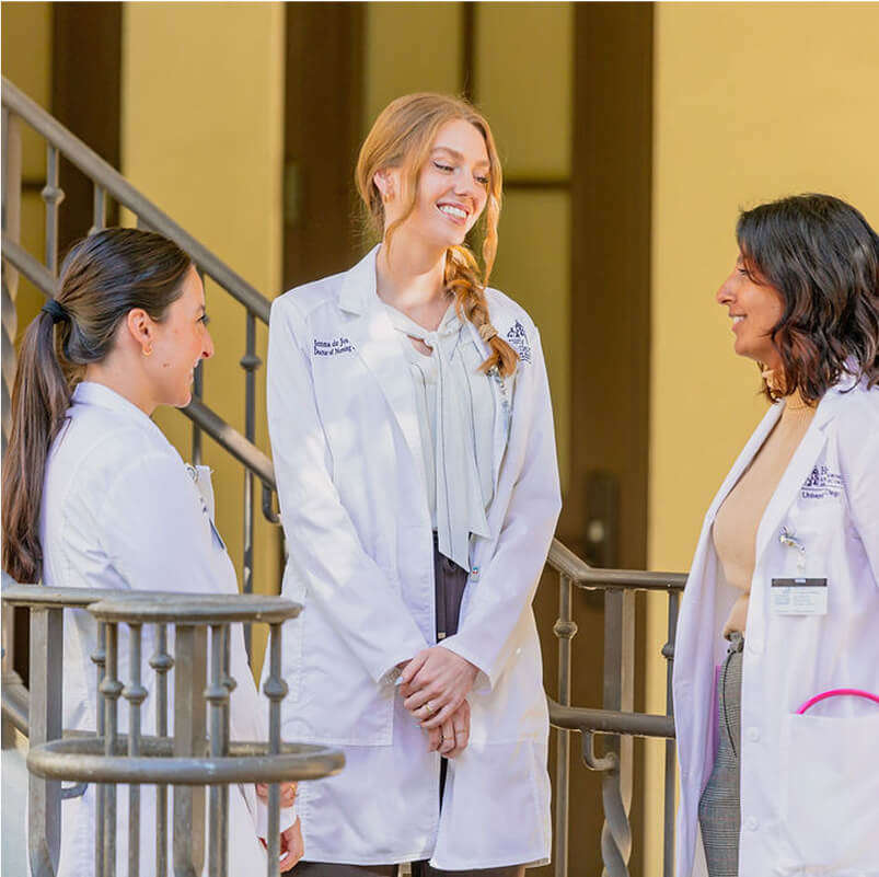 Three nursing students in white lab coats talking next to outdoor staircase 