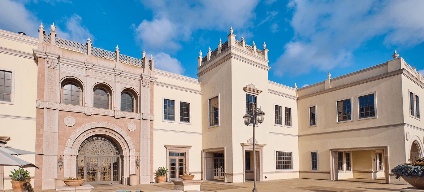 A cream-colored Spanish-style university building with an ornate tower and rounded archway doorway against a blue sky.
