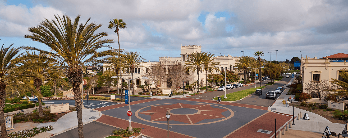 Aerial view of a palm-lined campus street with Spanish-style buildings and a decorative circular paved plaza in the foreground.