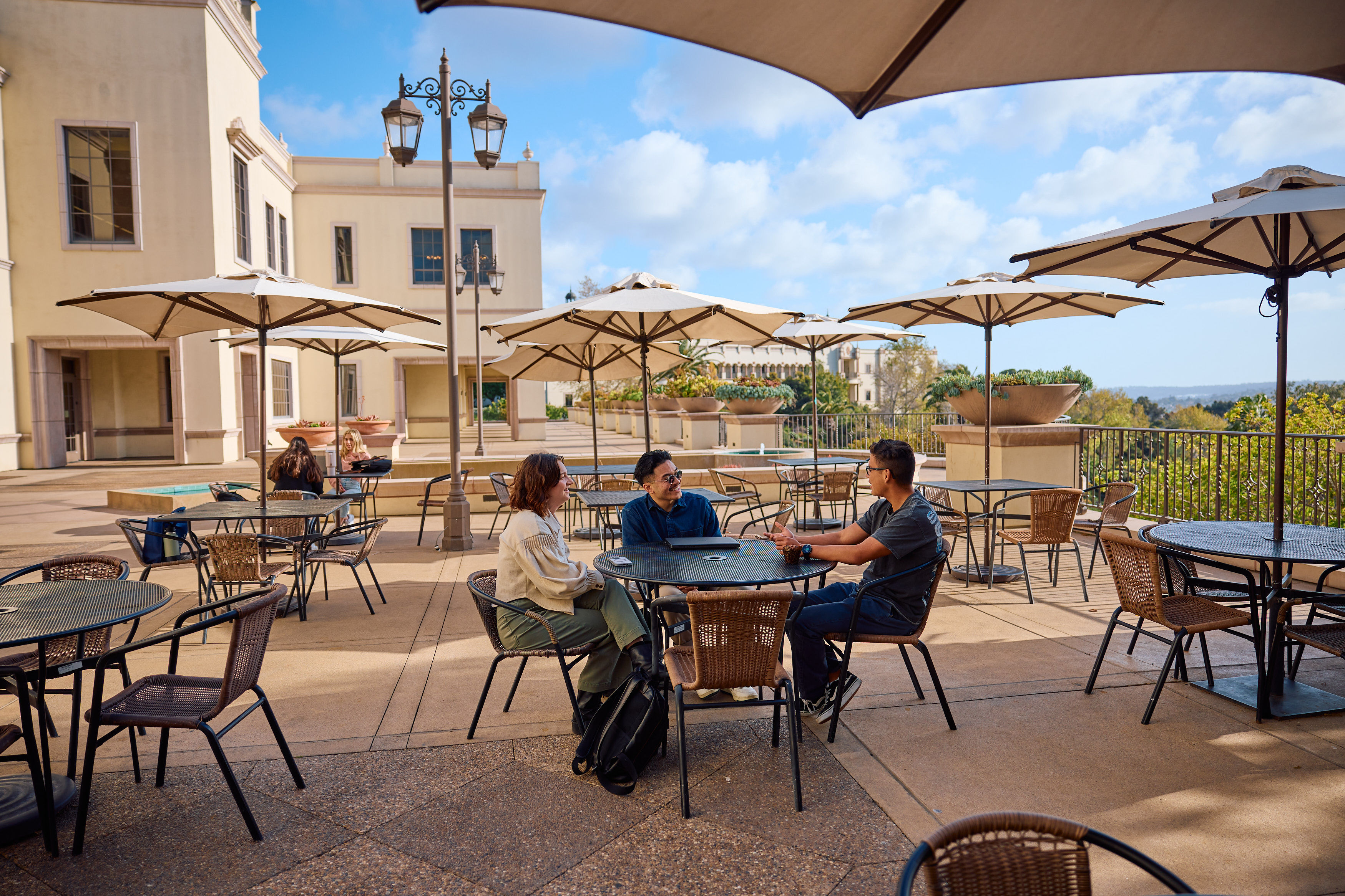 Three students conversing at a table on a sunny campus terrace with umbrellas and a view of the University of San Diego buildings and hillside.