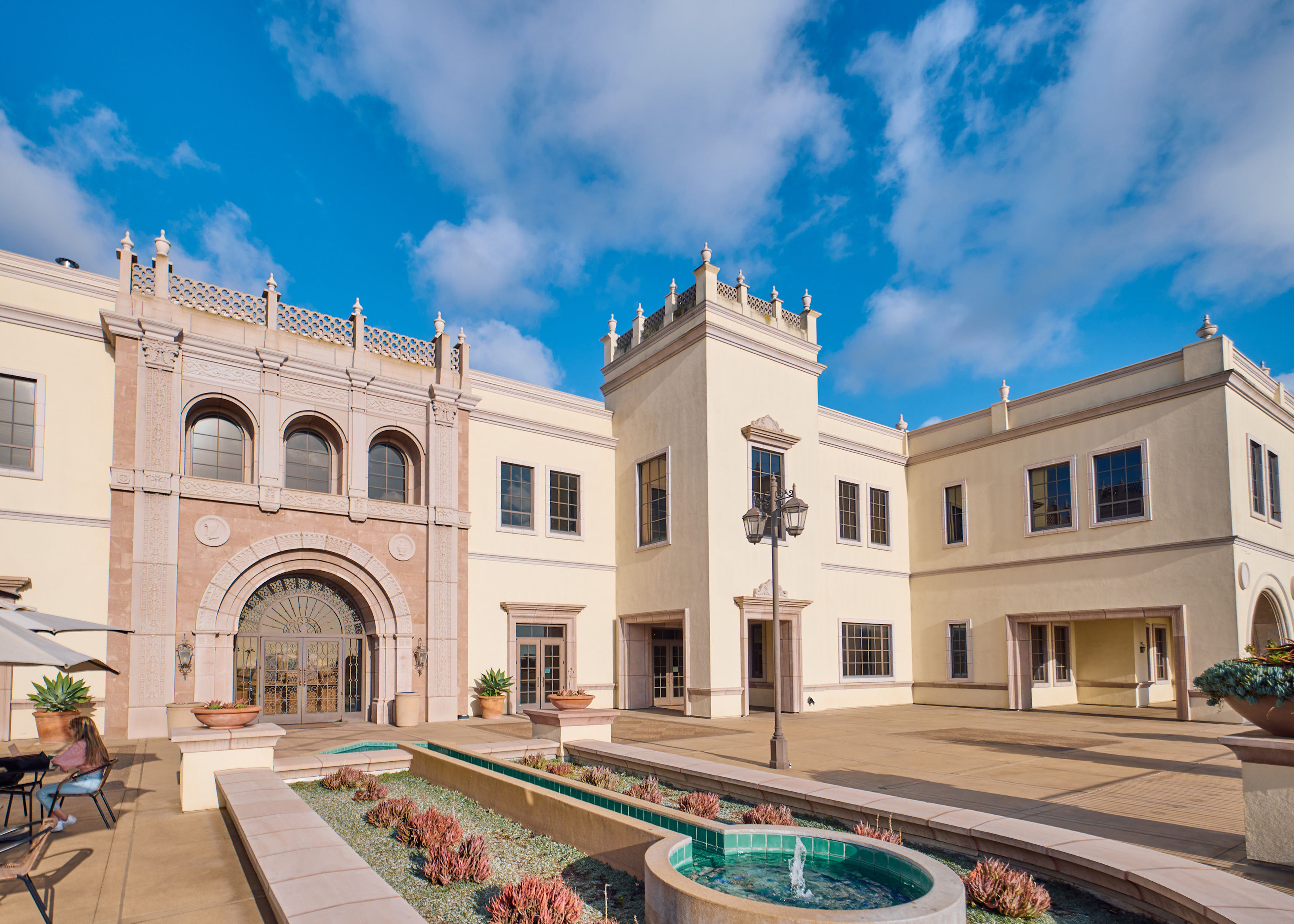 A cream-colored Spanish-style university building with an ornate tower and rounded archway doorway against a blue sky.
