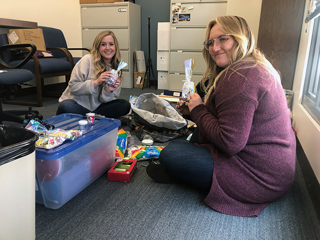 Two female with blonde hair students sitting on the office floor packing goodie bags. 