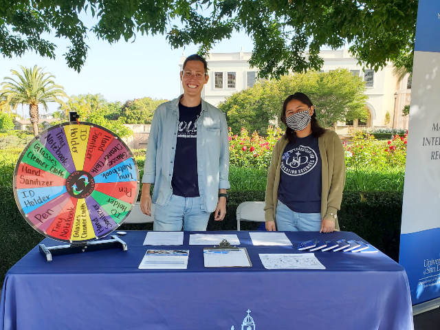 Two students, one male and one female, standing behind a table with a blue table cloth with a spining wheel.