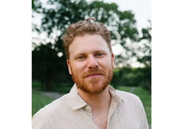This is a picture of a man with curly light brown hair and a white and green shirt against a green background. 