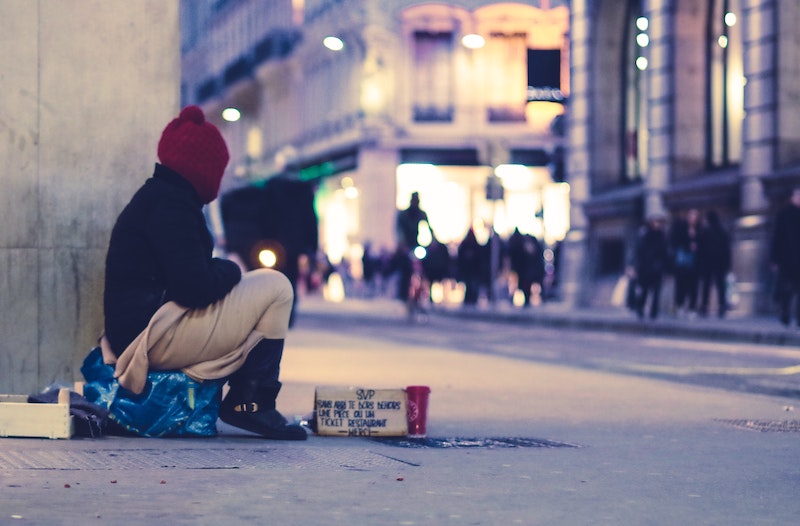 Person huddled up in sweater and beanie sits on a street corner with a sign asking for help