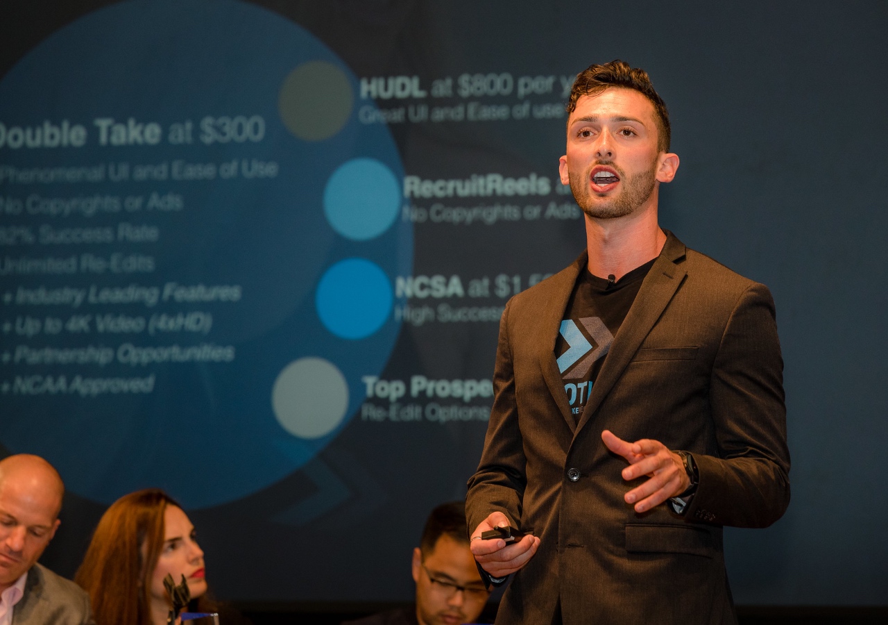 Student speaking on stage with a blue presentation behind him pitching a business idea