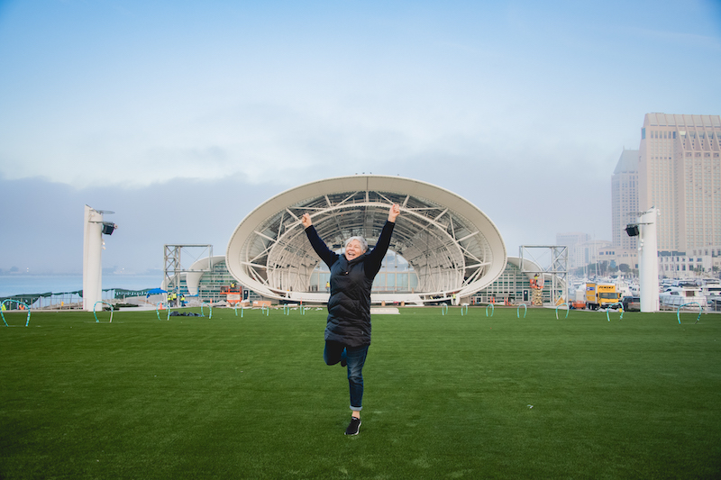 Katy McDonald jumps with her hands overhead in front of The Rady Shell at Jacobs Park