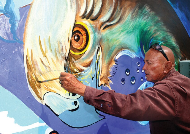 A photograph of Guillermo Aranda painting a large mural of a bird