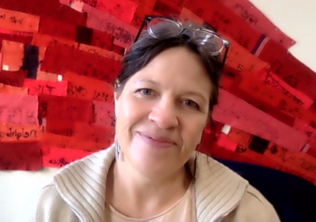 Marie is standing in front of a red fiber art piece, with her head tilted to the right, in a beige coat and glasses on her head.