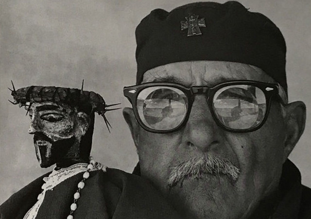 A grayscale photo of a man's face with glasses and a salt and pepper mustache. He is holding a figure of Jesus close to his face. 