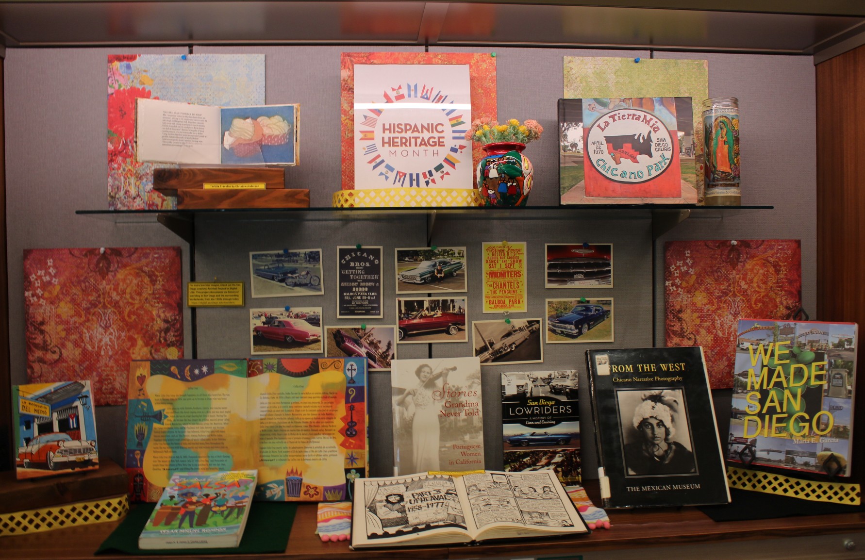 a glass case displaying a mural dedicated to Hispnaic Heritage Month, including photos of Hispanic artwork, photos of vintage cars, and a number of open books by and about Hispanic people