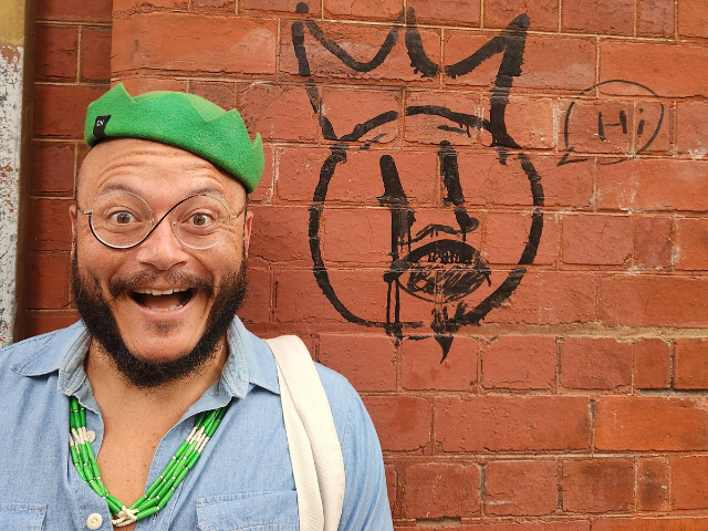 A picture of Dr. TJ Tallie in front of a brick wall with graffiti art on it.