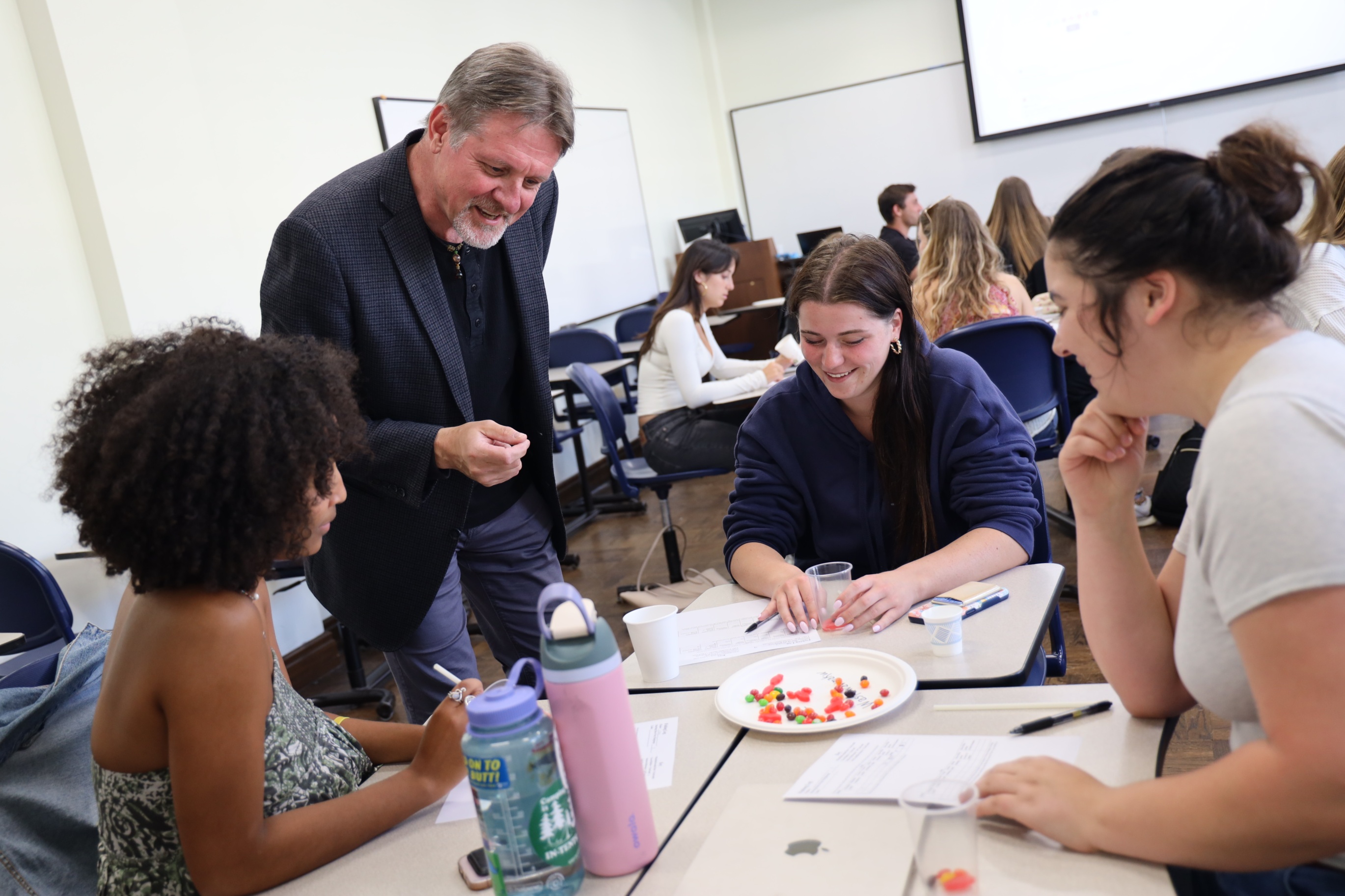 Dr. David Syring, USD Anthropology Chair, interacts with students during a classroom activity involving group discussion and colorful materials on a plate.
