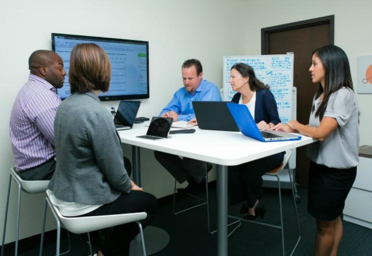 Group of diverse individuals around a table, with laptops, discussing research.