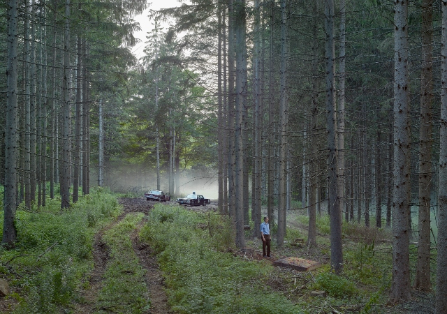 This is a photograph of a forest in fog. A tire track leads to two police cars in the background. To the right of the tracks, a person in a blue shirt stands, looking at a dirty mattress on the ground
