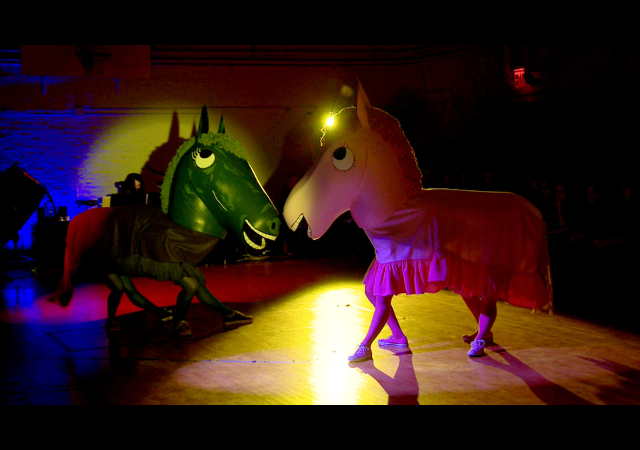 This is a picture of four people in costumes. The costumes are two horses, one green, one yellow. They are dancing on a stage.