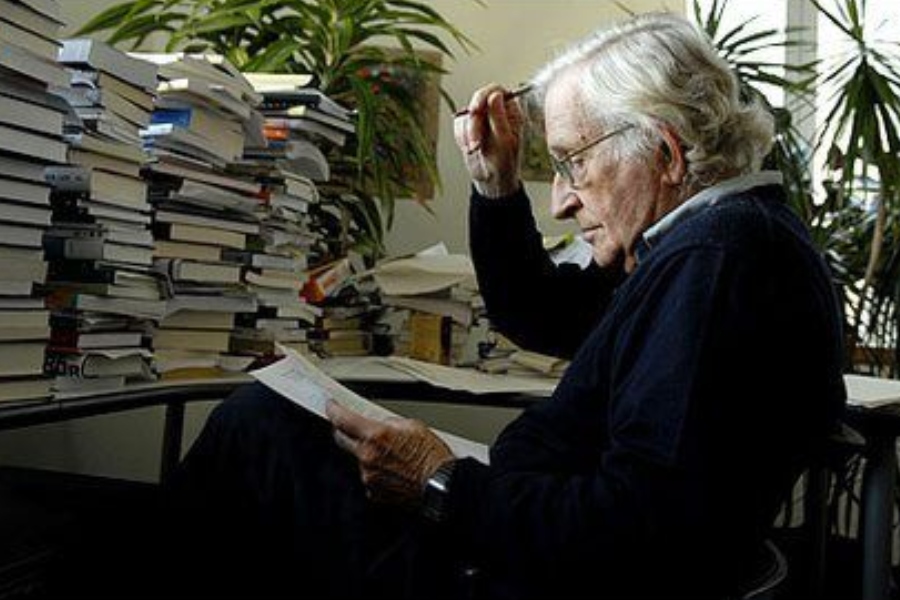 Noam sitting in a chair, reading a book, in front of a desk piled high with books. There is a potted plant in the background.