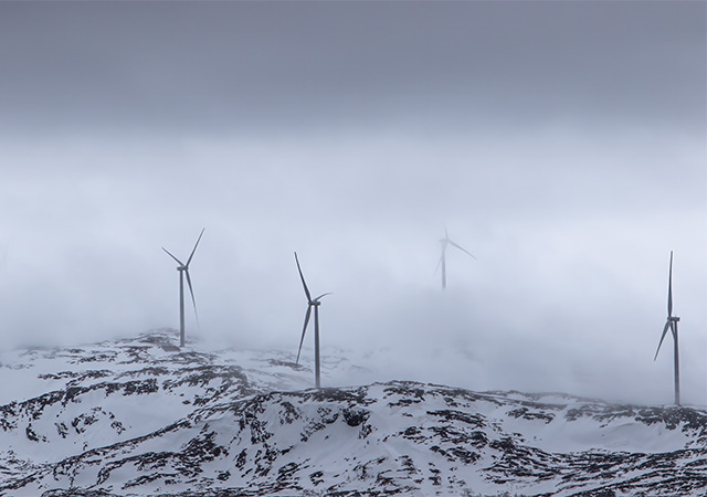 A picture of a rocky outcrop covered in snow. There are four wind turbines enshrouded in fog.