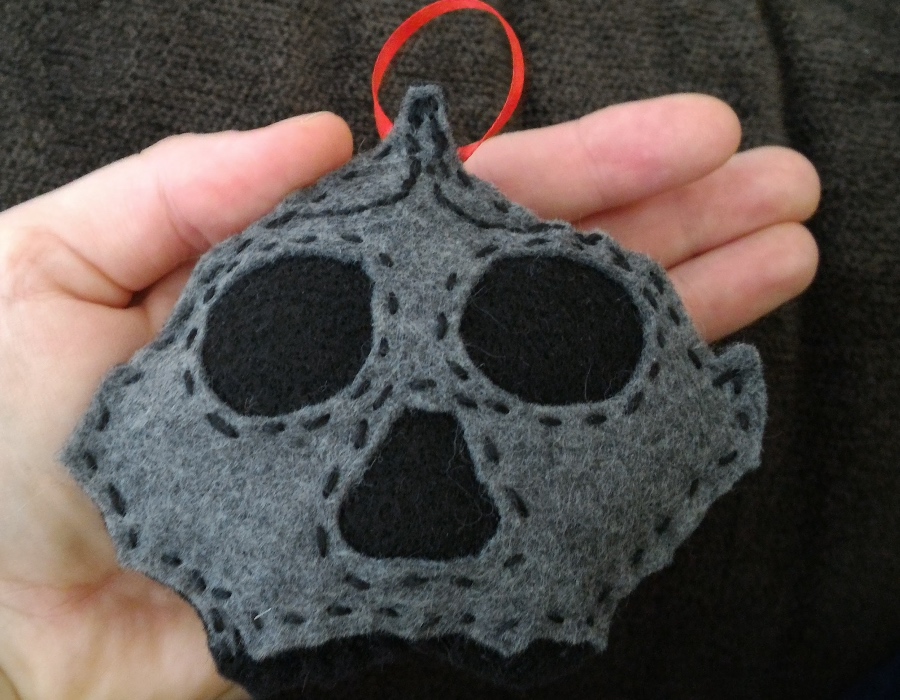 open palm holding a grey, hand sewn skull shaped ornament with a red ribbon hanger