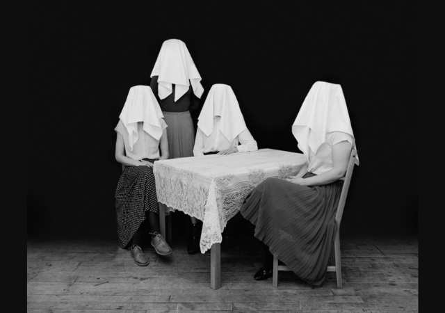 This is a black and white picture of four women sitting at a table with white clothes over their heads. 