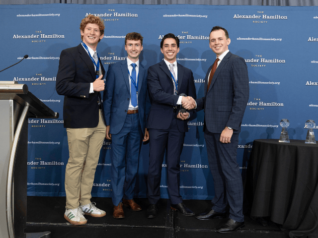 Four individuals in formal attire stand in front of a branded backdrop for The Alexander Hamilton Society. Two shake hands in the center, with a podium on the left and a table displaying glass awards on the right, suggesting a ceremonial or recognition event.